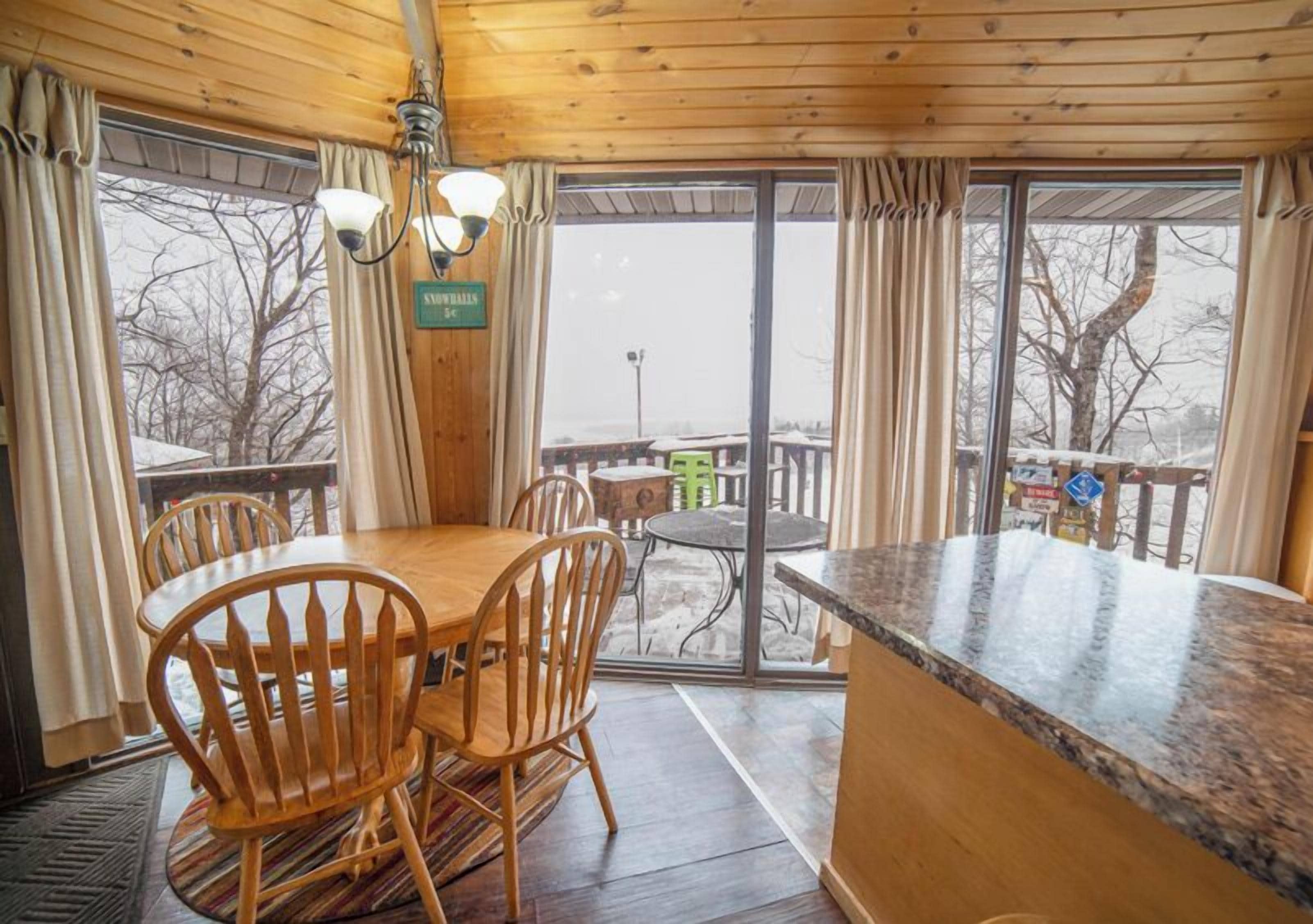 A wooden interior with a dining table and chairs, a kitchen counter, and large windows with curtains.