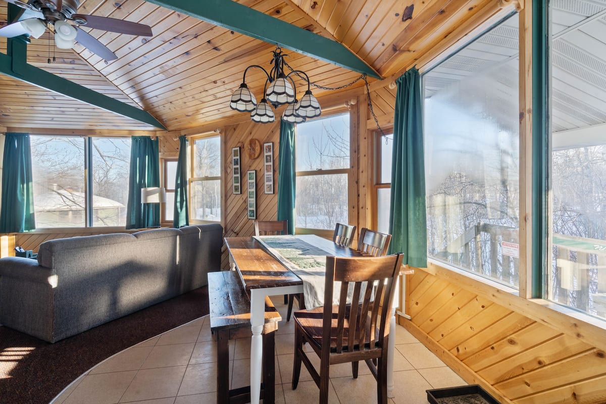 A cozy cabin interior with a wooden ceiling, large windows, a dining table, and a sofa.
