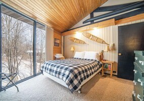 A bedroom with a large window, a bed with a checkered bedspread, a bedside table with a lamp, and a bookshelf.