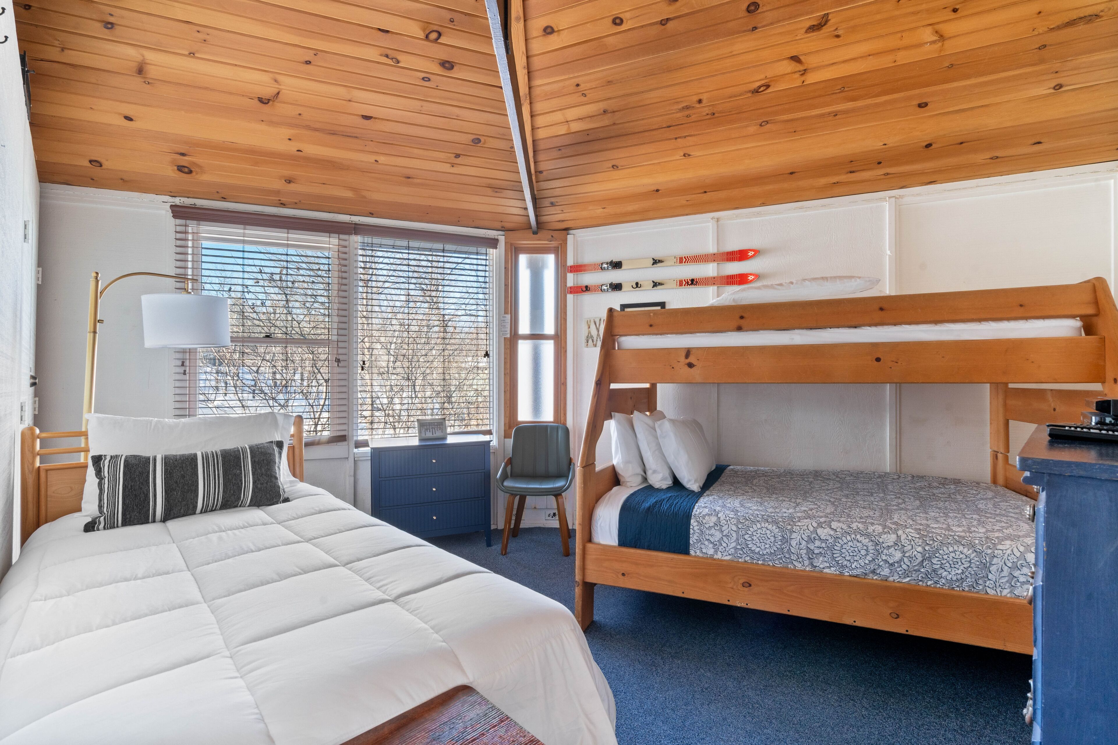 A bedroom with a bunk bed, a desk with a lamp, a chair, and a window with blinds.