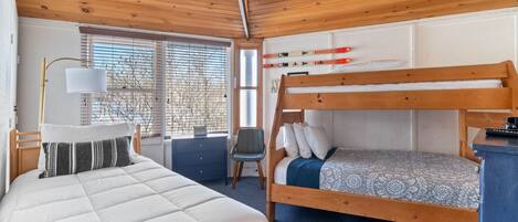 A bedroom with a bunk bed, a desk with a lamp, a chair, and a window with blinds.