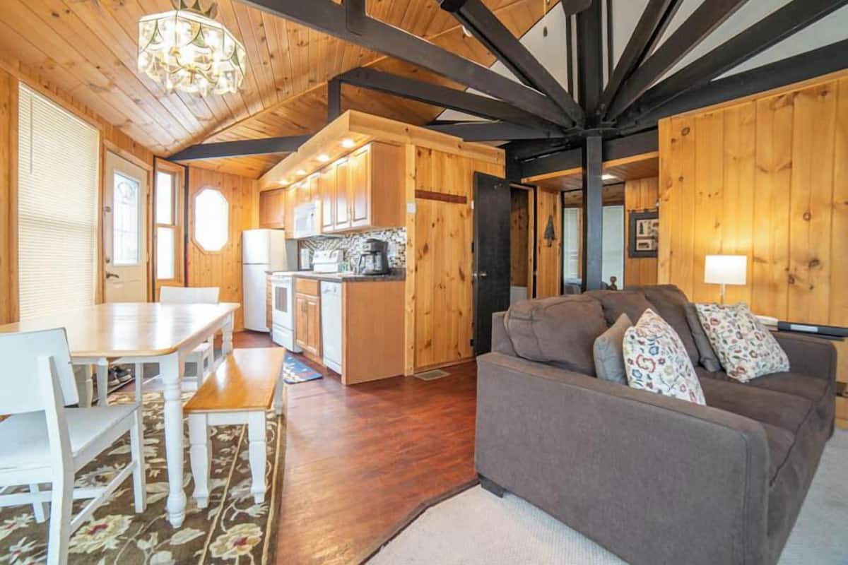 A cozy cabin interior with a wooden dining area, a kitchenette, and a living room with a sofa and pillows.