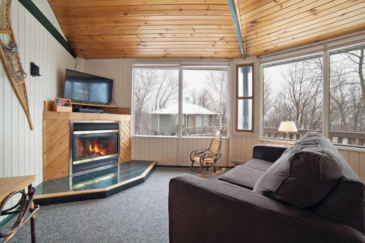 A cozy living room with a fireplace, a television, and a large window offering a view of the outdoors.