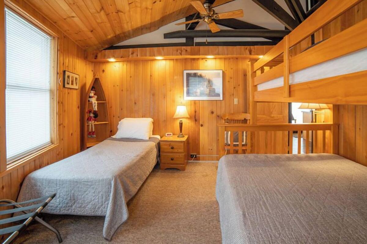 A wooden cabin bedroom with two beds, a ceiling fan, and a window with blinds.