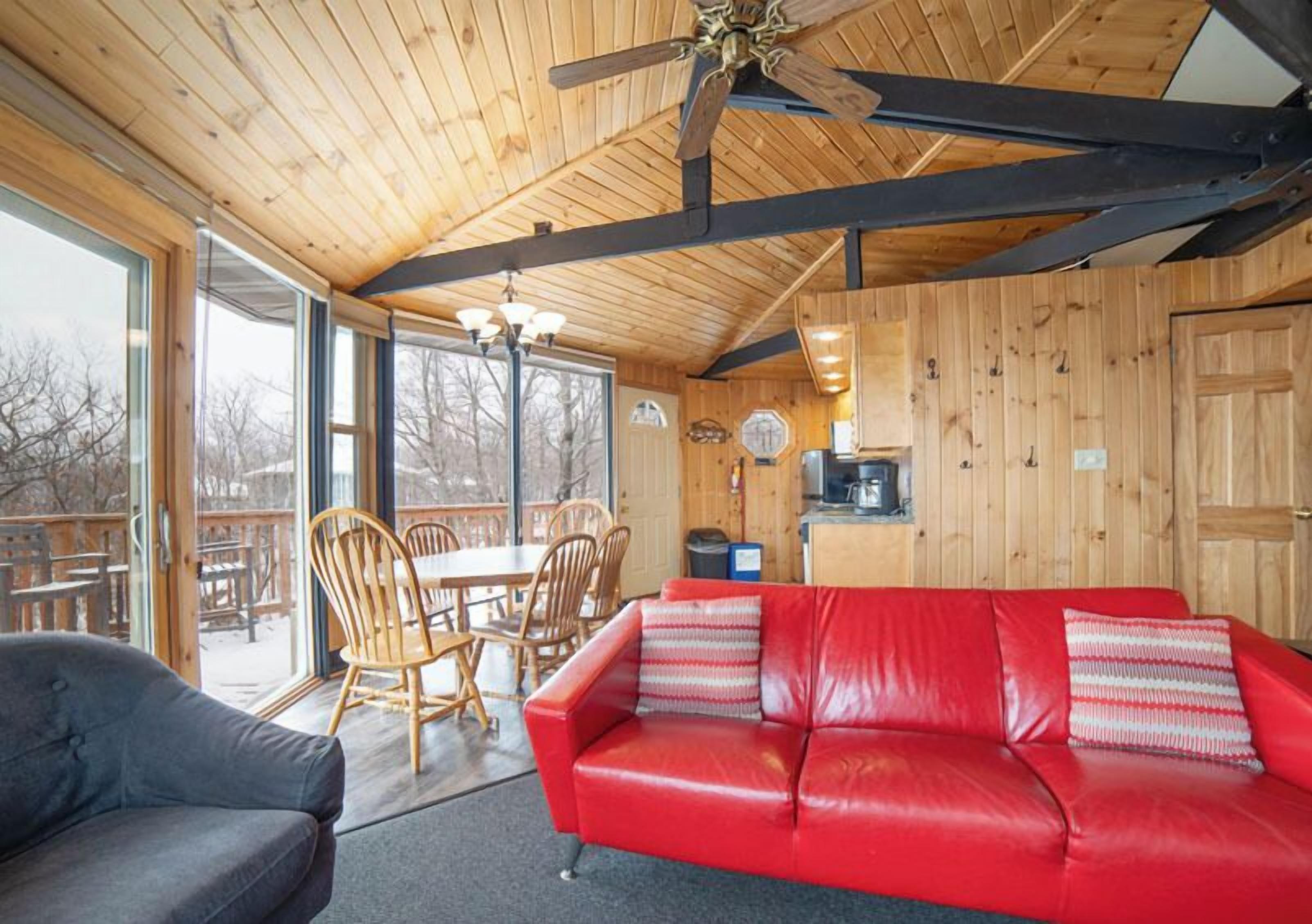 Interior de una cabaña de madera con un sofá de cuero rojo, una mesa de comedor de madera, sillas y un ventilador de techo.