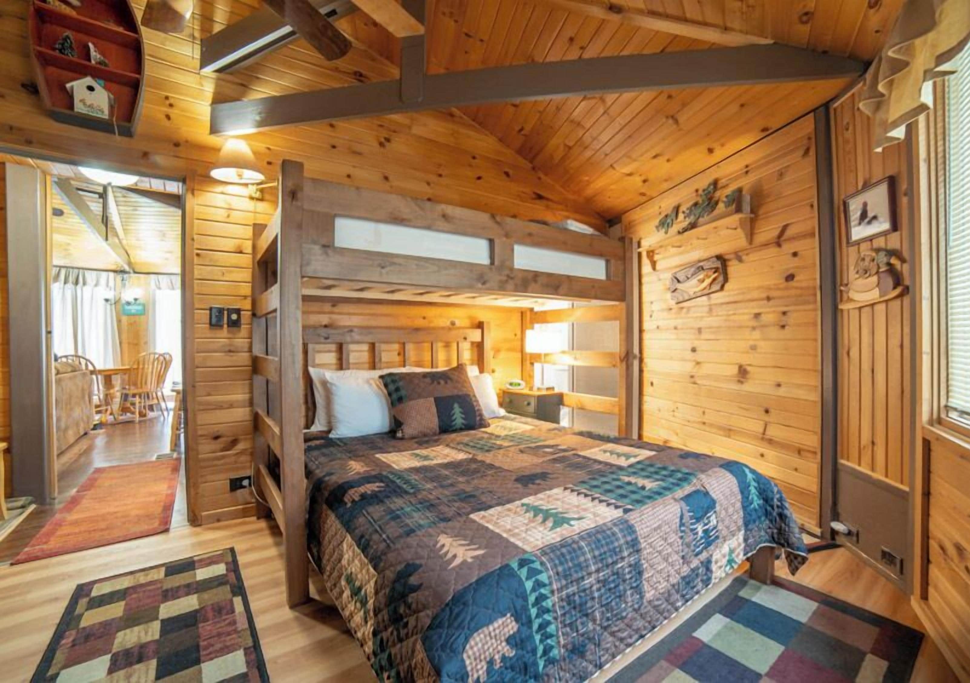 A cozy wooden cabin room with a large bed, a wooden headboard, a window, and a door leading to another room.