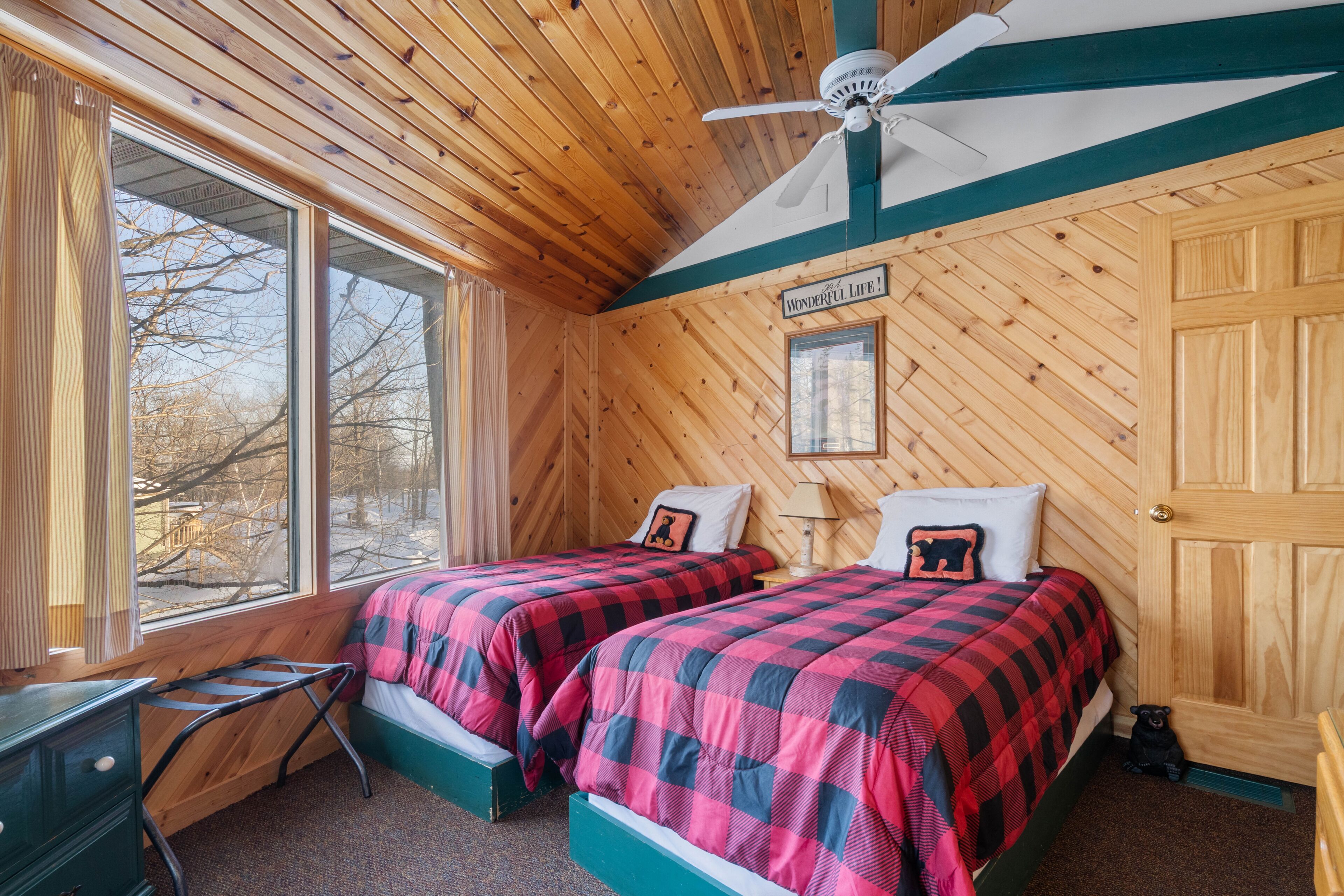 Habitació d'una cabana de fusta amb dos llits, un ventilador de sostre i una finestra amb vista a arbres.