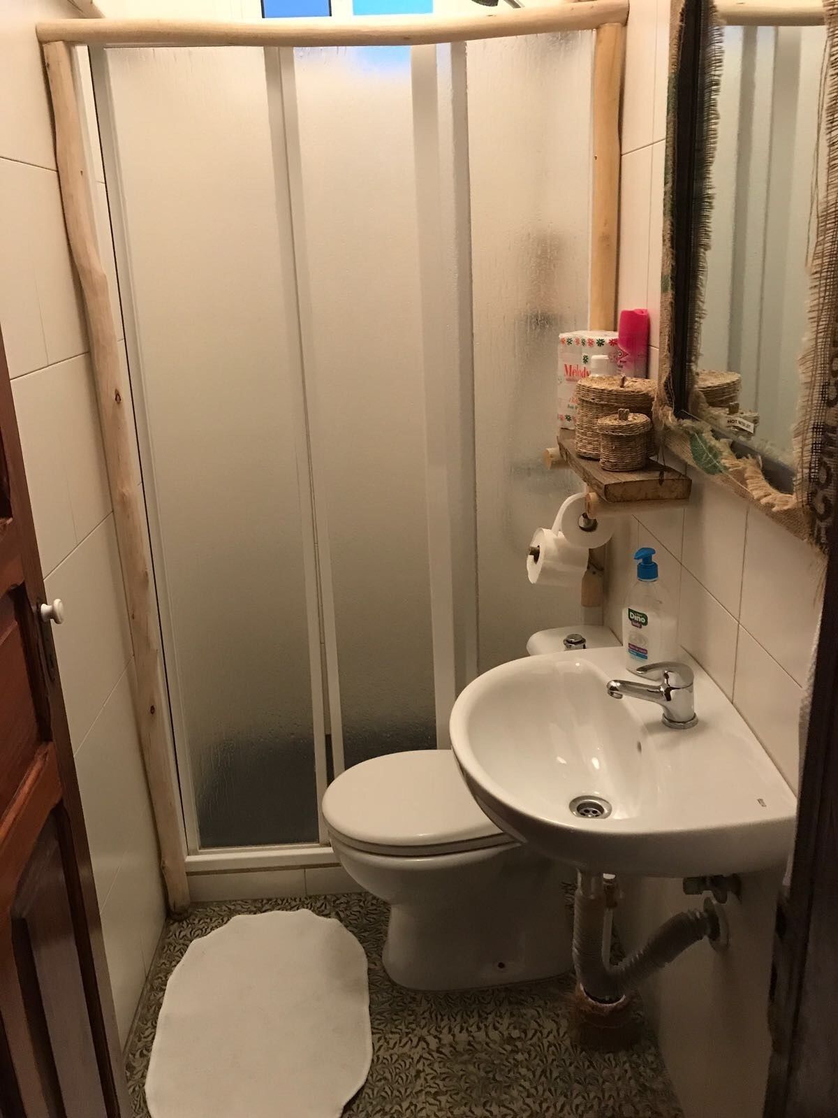A small bathroom with a toilet, sink, and shower. There is a towel on the floor and toiletries on the sink.