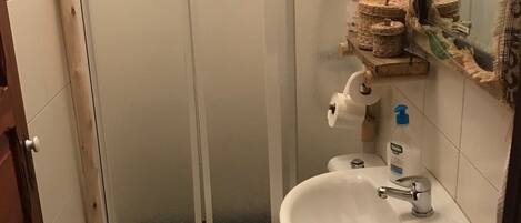A small bathroom with a toilet, sink, and shower. There is a towel on the floor and toiletries on the sink.