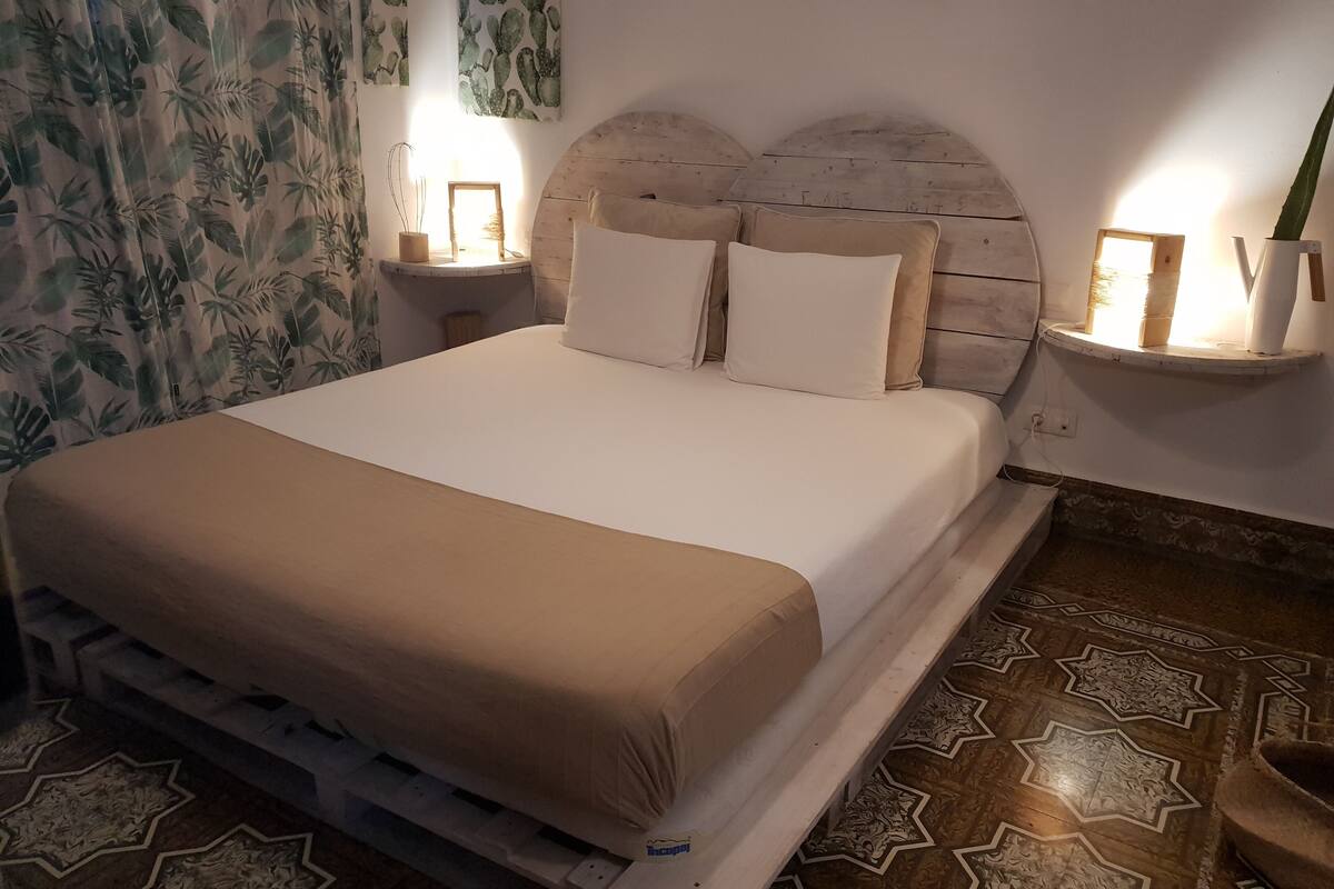 Een net opgemaakt bed met een houten hoofdbord, een nachtkastje met een lamp en een wandplank met een plantje.
