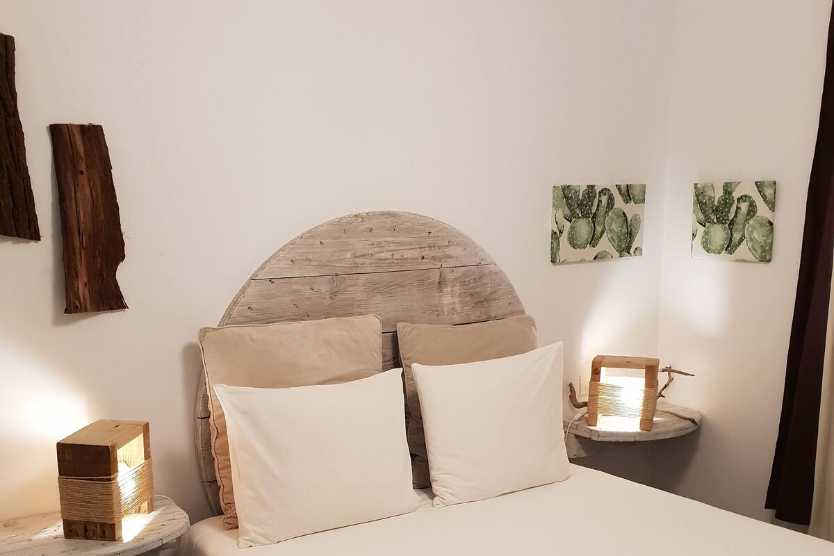 Een netjes opgemaakt bed met een houten hoofdbord, twee nachtkastjes met lampen, en wanddecoratie met cactussen.