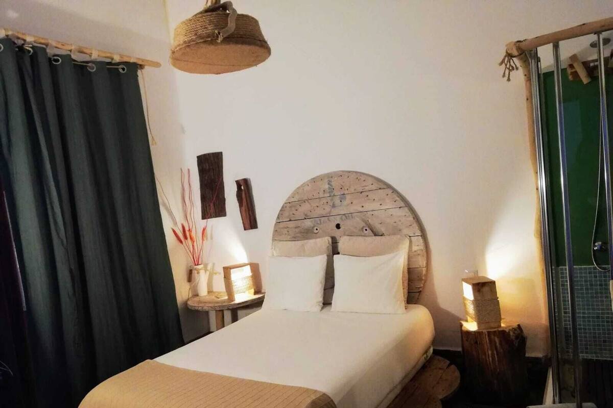 Een net opgemaakt bed met houten hoofdeinde, een nachtkastje met lamp, en een gordijn aan de linkerkant.