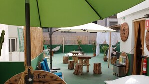Un espacio al aire libre rústico con mesas y bancos de madera, rodeado de follaje y un muro cubierto de plantas.