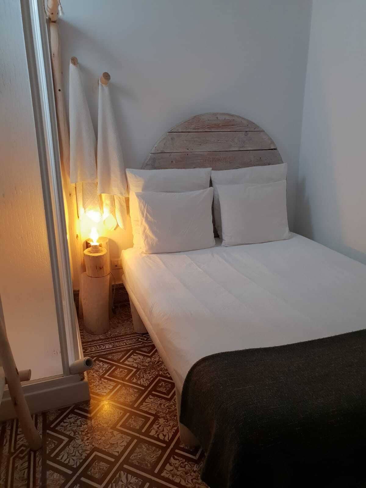 Un dormitorio con cama, ropa de cama blanca, cabecero de madera, mesita de noche con lámpara y baldosas de suelo estampadas.