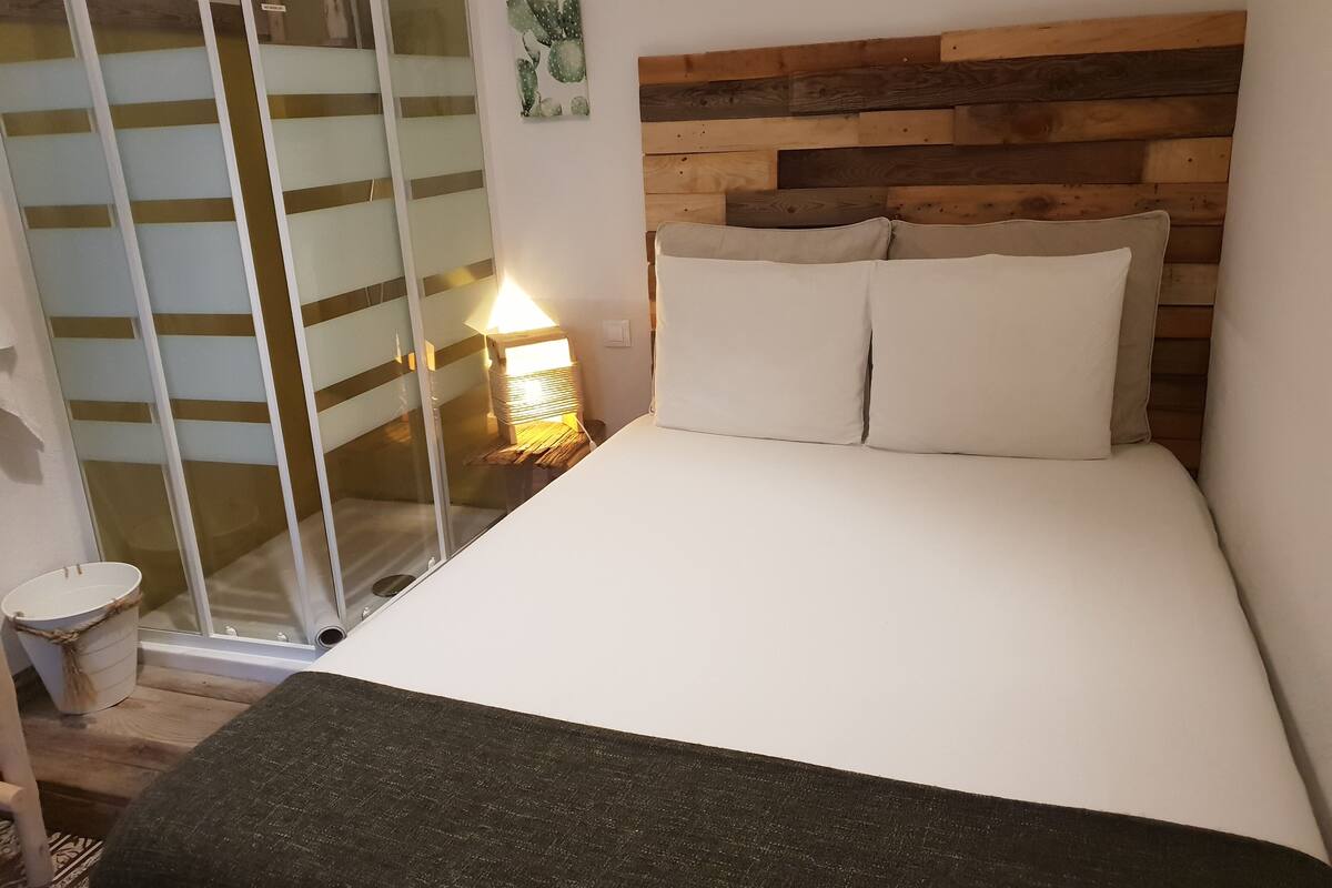Een slaapkamer met een houten hoofdbord, een bed met witte beddengoed, een bedlampje en een glazen deur naar een douchegedeelte.
