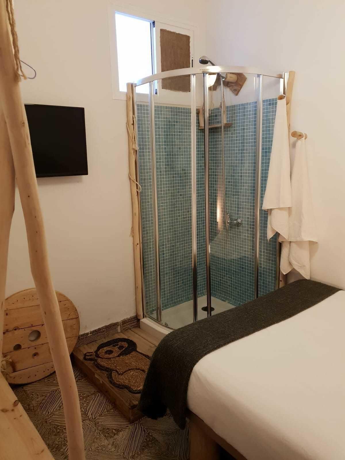 Une petite chambre accueillante, dotée d’un lit, d’une douche avec des portes en verre, d’un plancher en bois et d’un téléviseur fixé au mur.