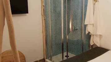 Une petite chambre accueillante, dotée d’un lit, d’une douche avec des portes en verre, d’un plancher en bois et d’un téléviseur fixé au mur.