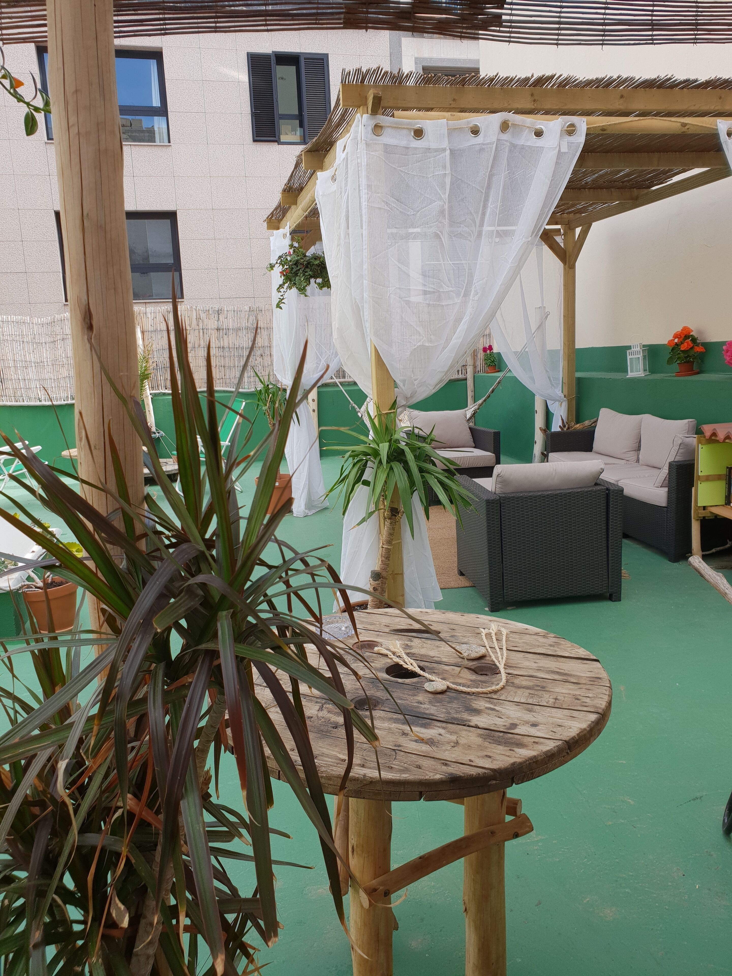 Een overdekt terras met een houten tafel, rieten meubels en potplanten.