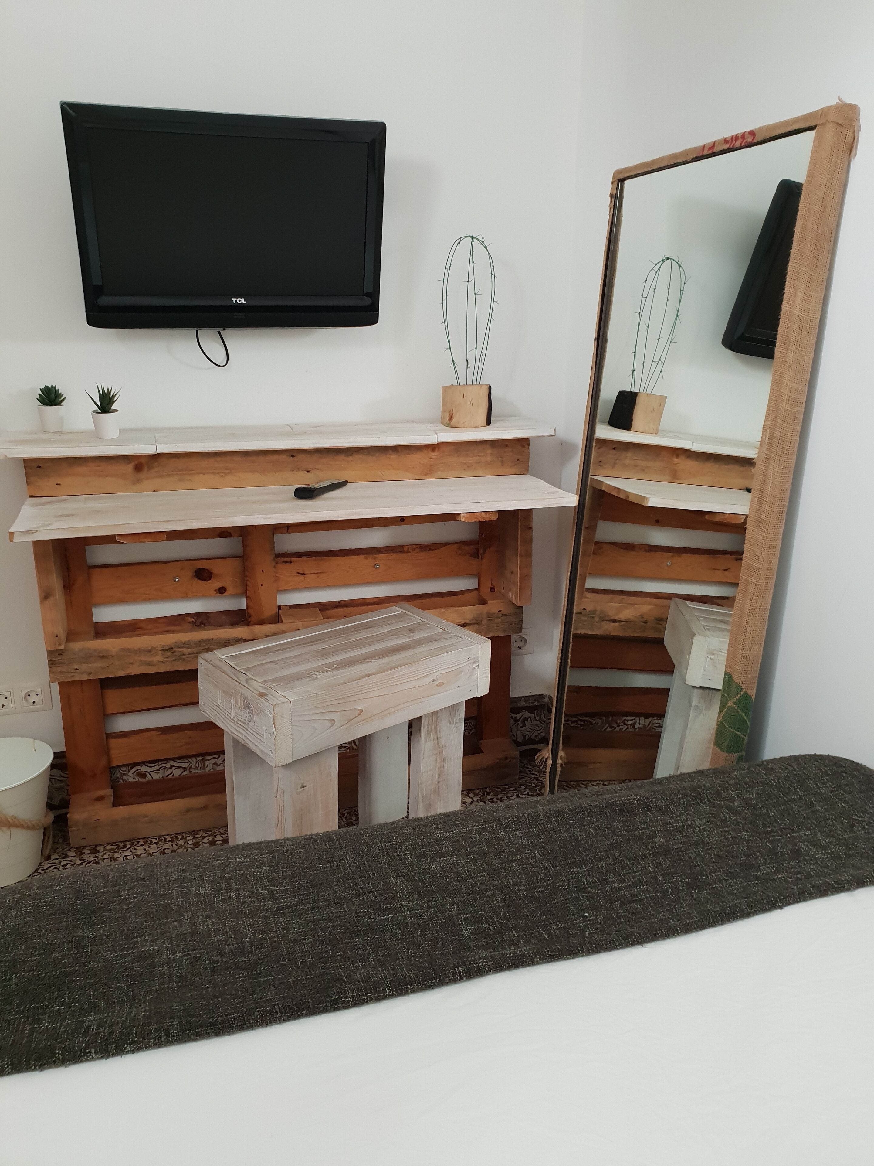 Una habitación con un banco de madera, una pequeña mesa, un televisor montado en la pared y un espejo con marco de madera.