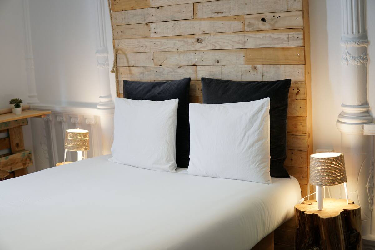 Een bed met witte lakens, twee zwarte kussens en twee nachtlampjes met gevlochten kapjes.
