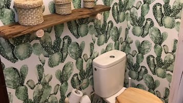 Een badkamer met cactuspapier, een houten plank met manden, een toilet met houten zitting en een emmer op de vloer.