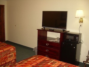 A hotel room with a bed, a wooden dresser with a television and a microwave, and a lamp on the wall.
