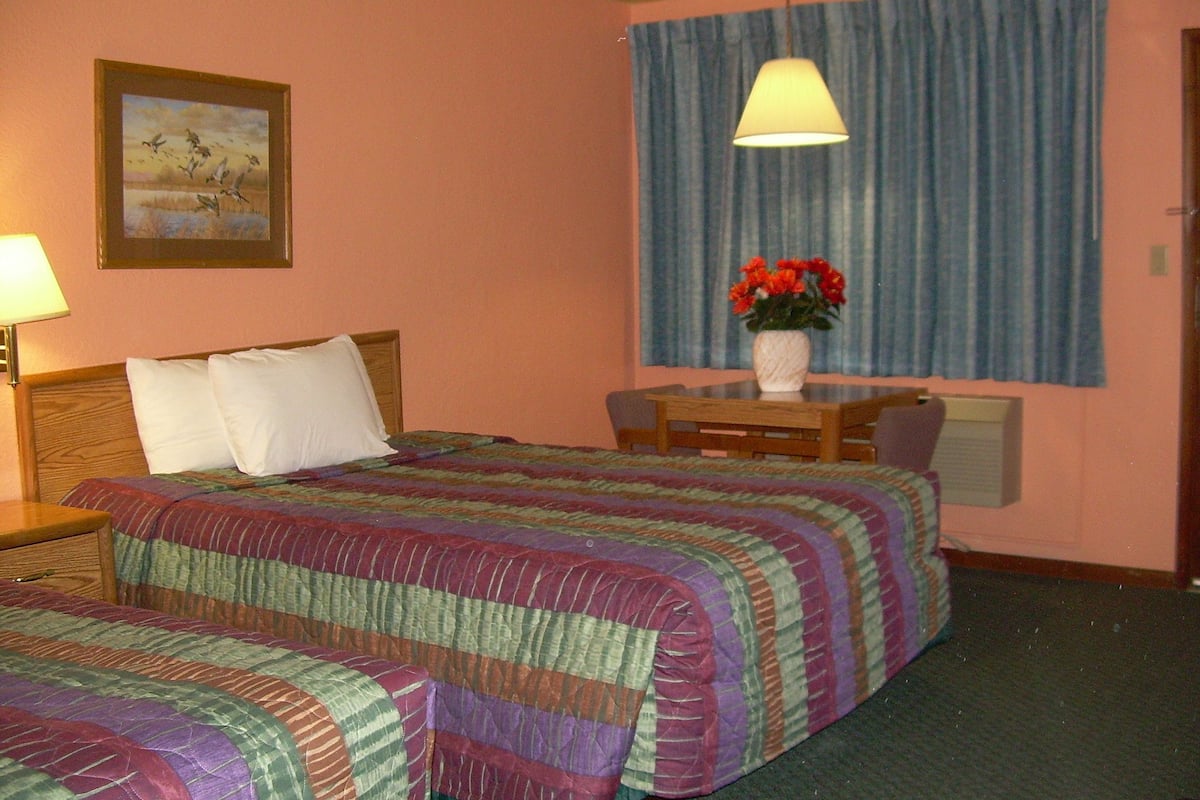 A hotel room with a bed, a desk, and a chair. There is a painting on the wall and a window with curtains.