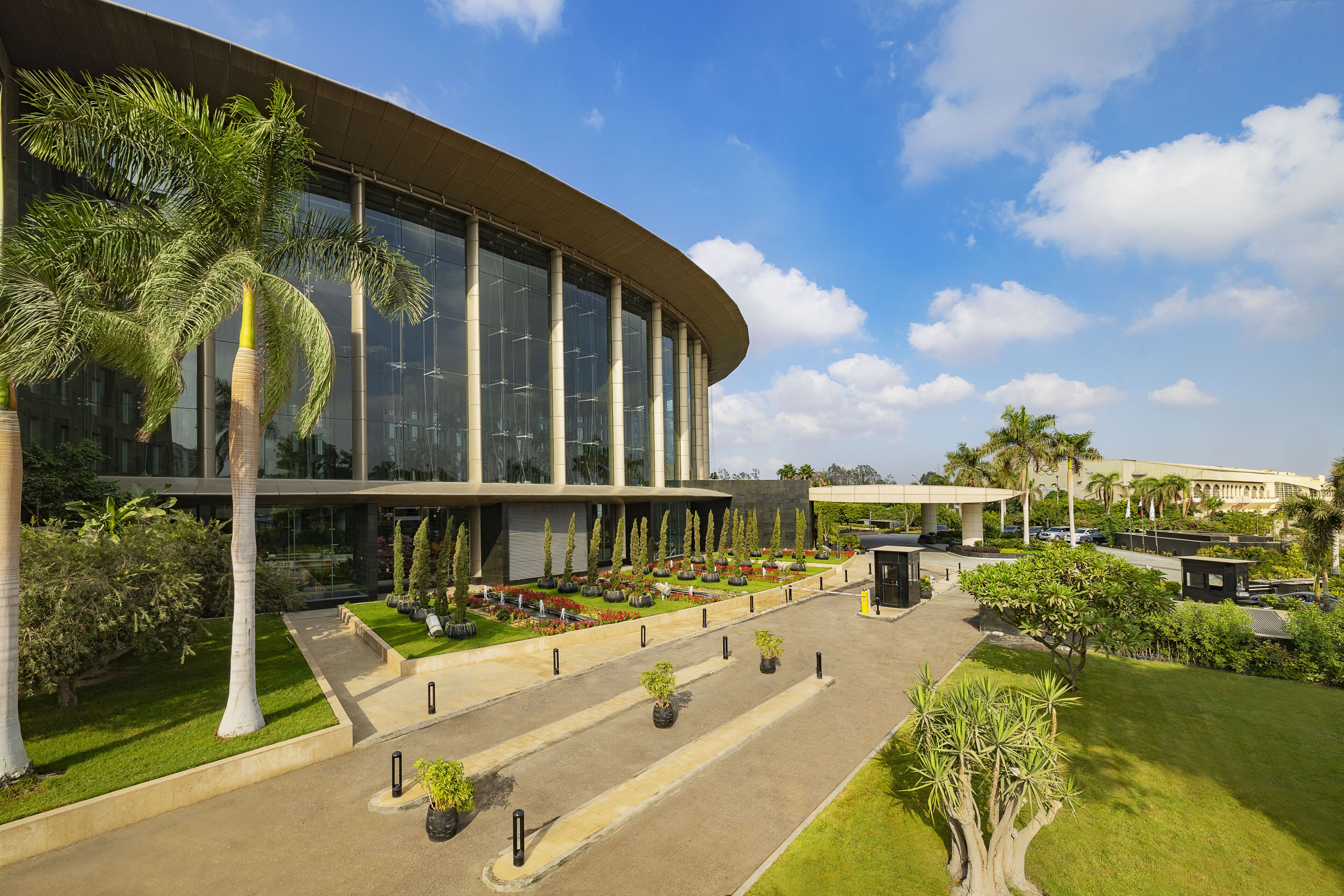 A modern building with a curved facade, large windows, and a well-maintained garden with palm trees and shrubs.