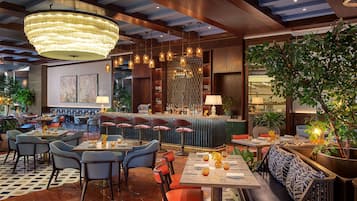 Un restaurante moderno con zona de bar, diversas opciones de asientos, incluyendo mesas y cabinas, y una gran lámpara de araña.