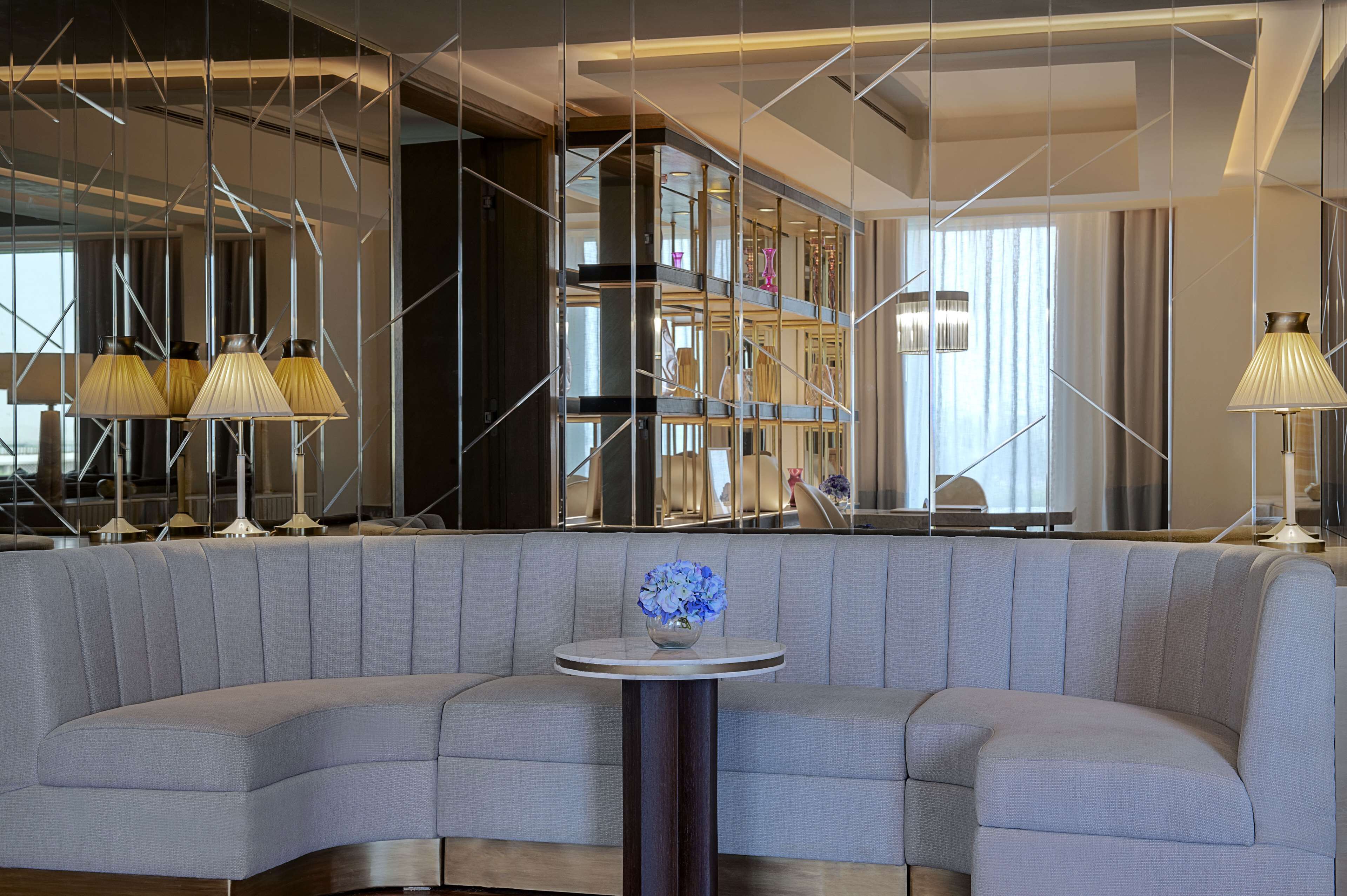 A modern hotel lobby with a curved gray sofa, a round table with a vase of flowers, and a large bookshelf.