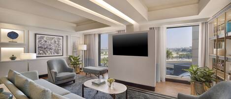 A modern hotel room with a large flat-screen TV, comfortable seating, a patterned rug, and a city view through large windows.
