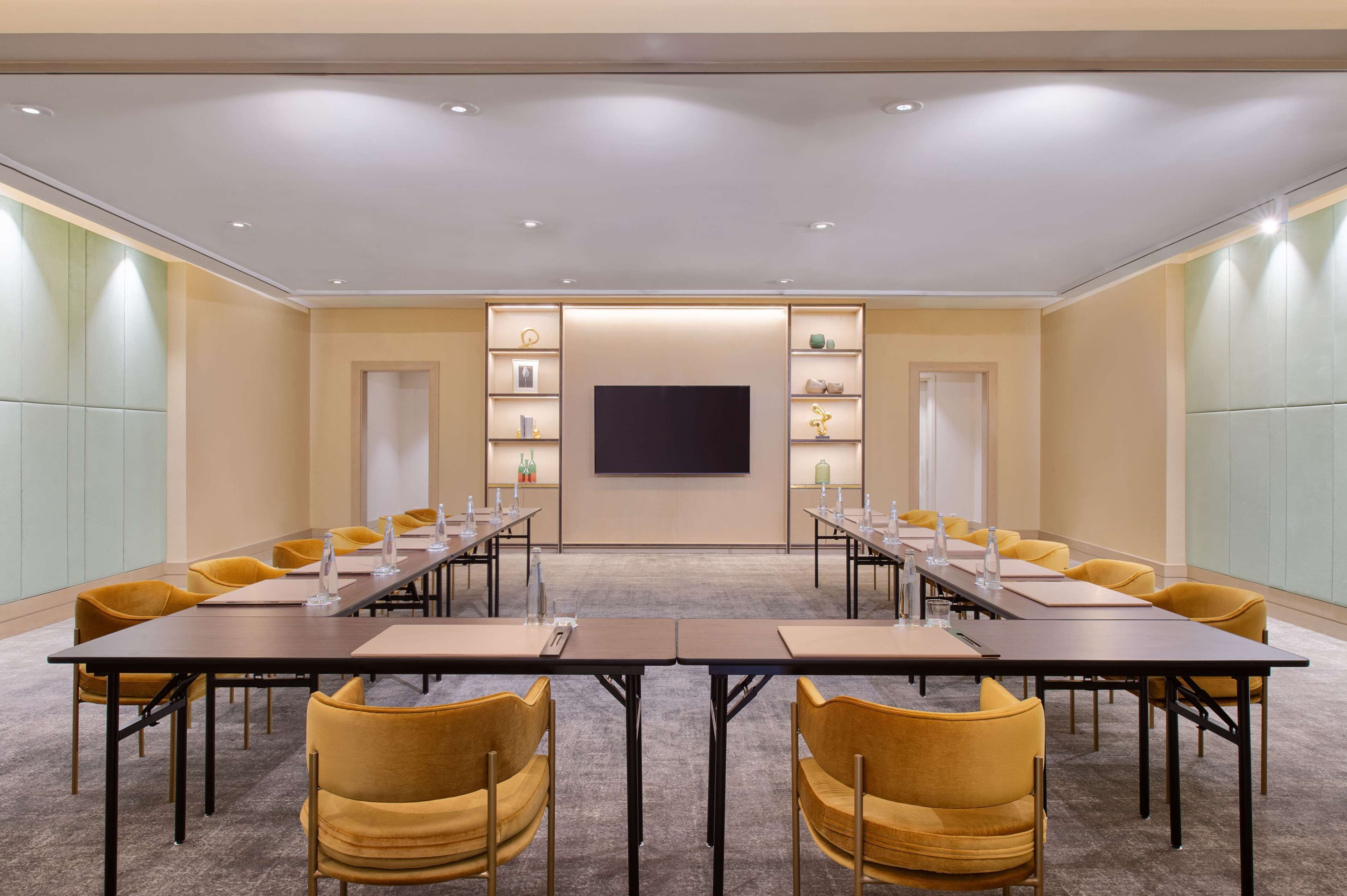 A conference room with a long table, yellow chairs, a flat-screen TV, and shelves with various items.