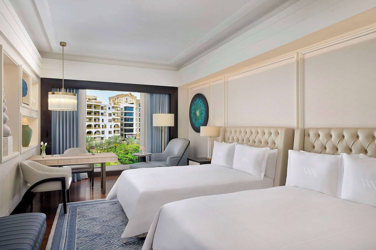 A hotel room with two beds, a desk, a chair, and a view of a building.
