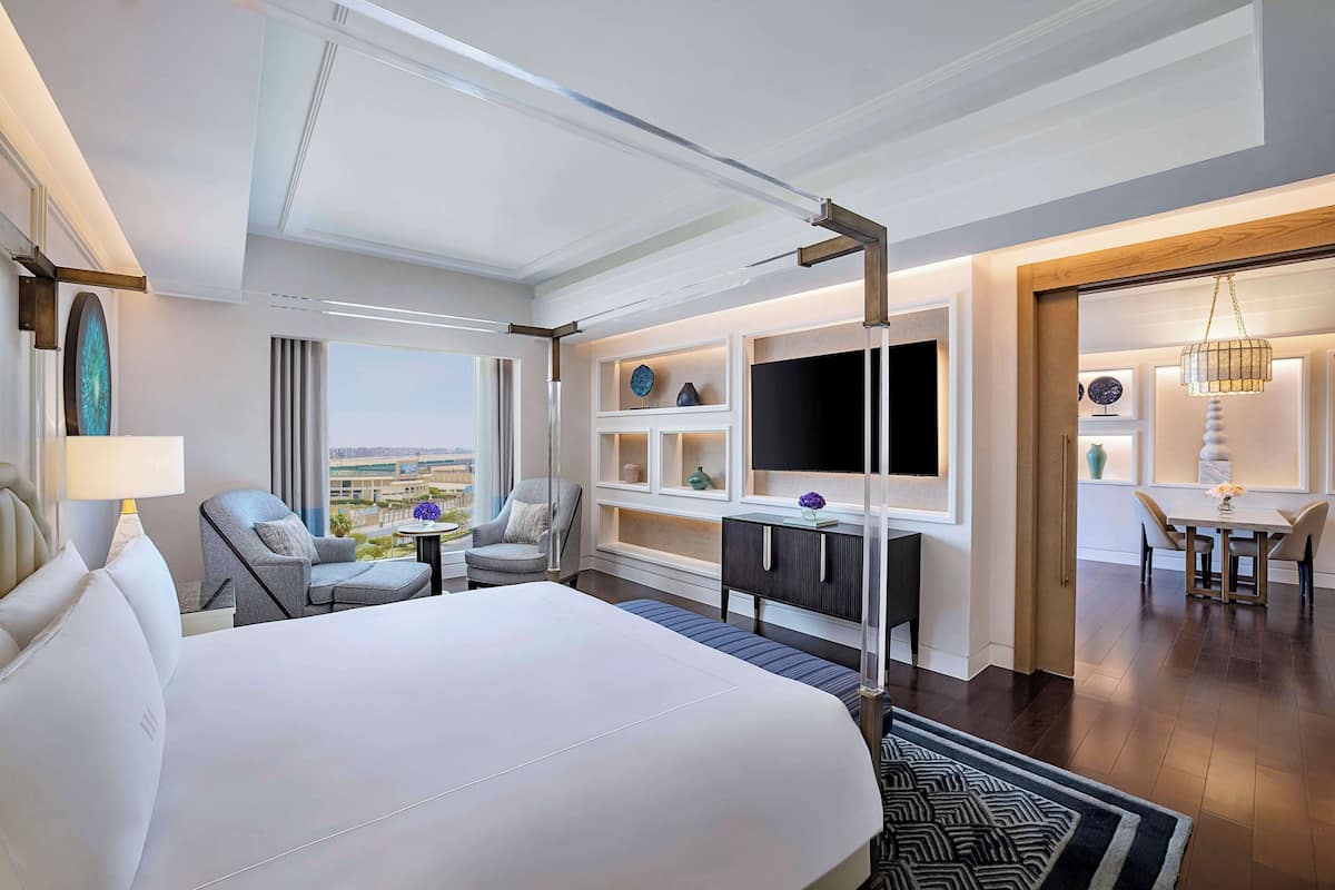 A modern hotel room with a large bed, a desk, a TV, and a dining area with a view.