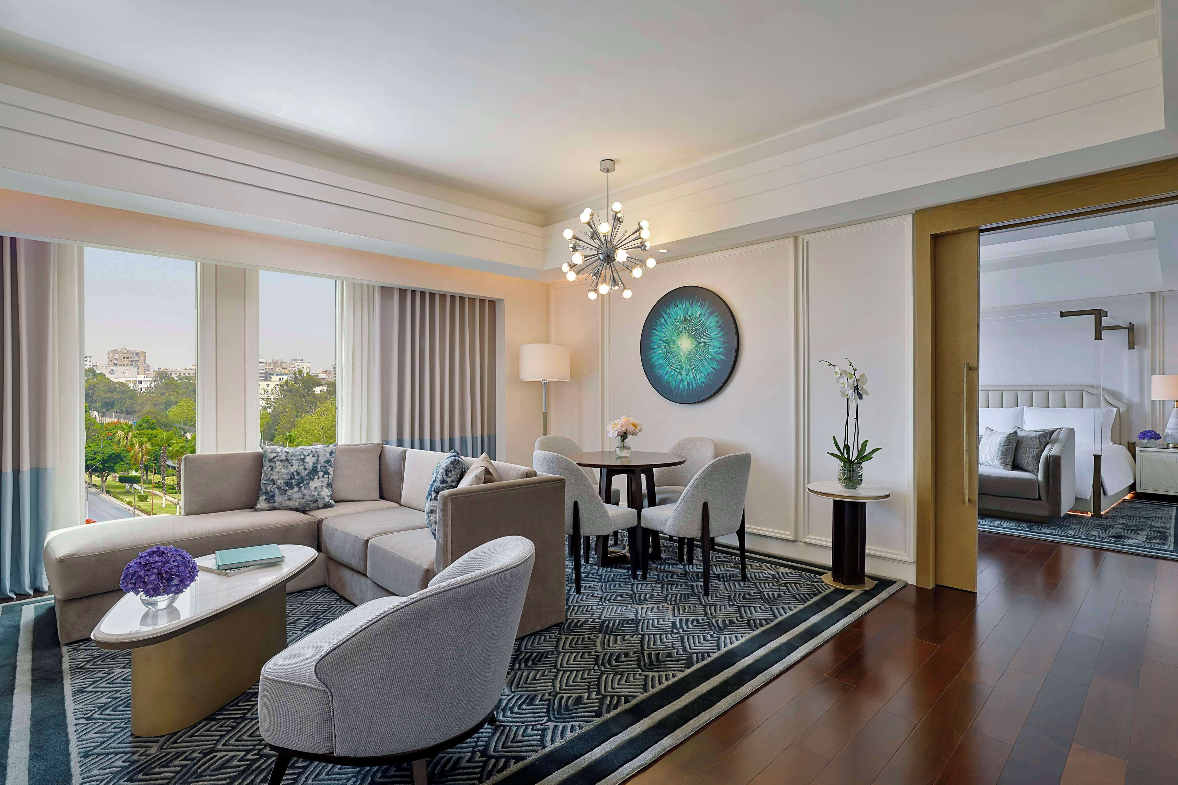 A modern hotel room with a large window, a sofa, a coffee table, a dining area, and a view of the cityscape.