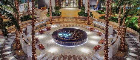 A spacious lobby with a central circular seating area, palm trees, and a geometric floor pattern.