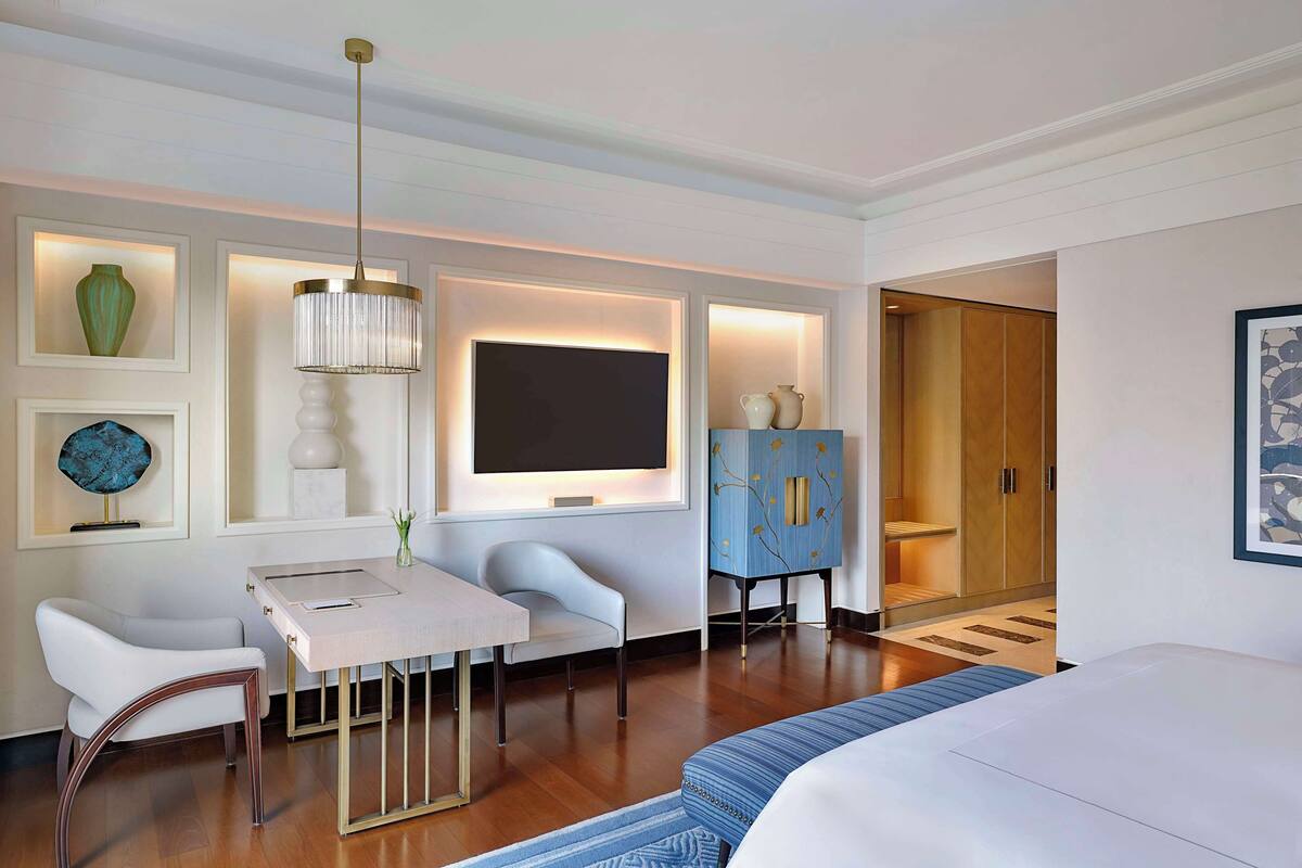 A hotel room with a bed, a desk with a chair, a TV, a wardrobe, and a decorative vase.