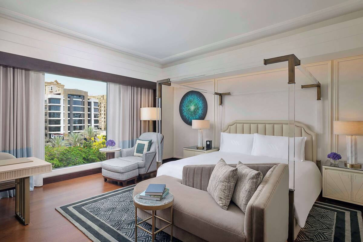 A hotel room with a large bed, a sofa, a chair, a small table, and a view of buildings and greenery outside.