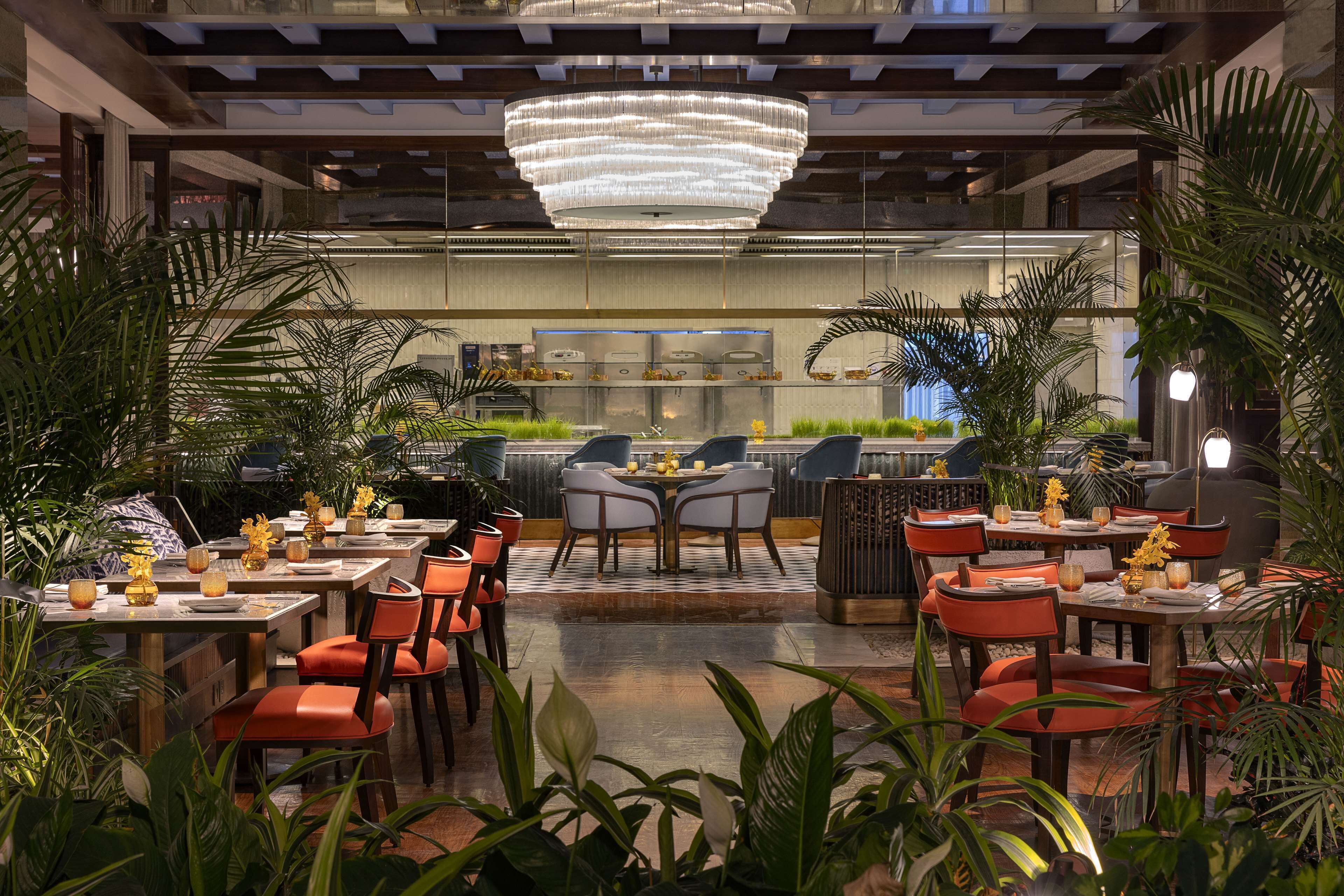 A restaurant with multiple dining tables and chairs, a large chandelier, and various plants.