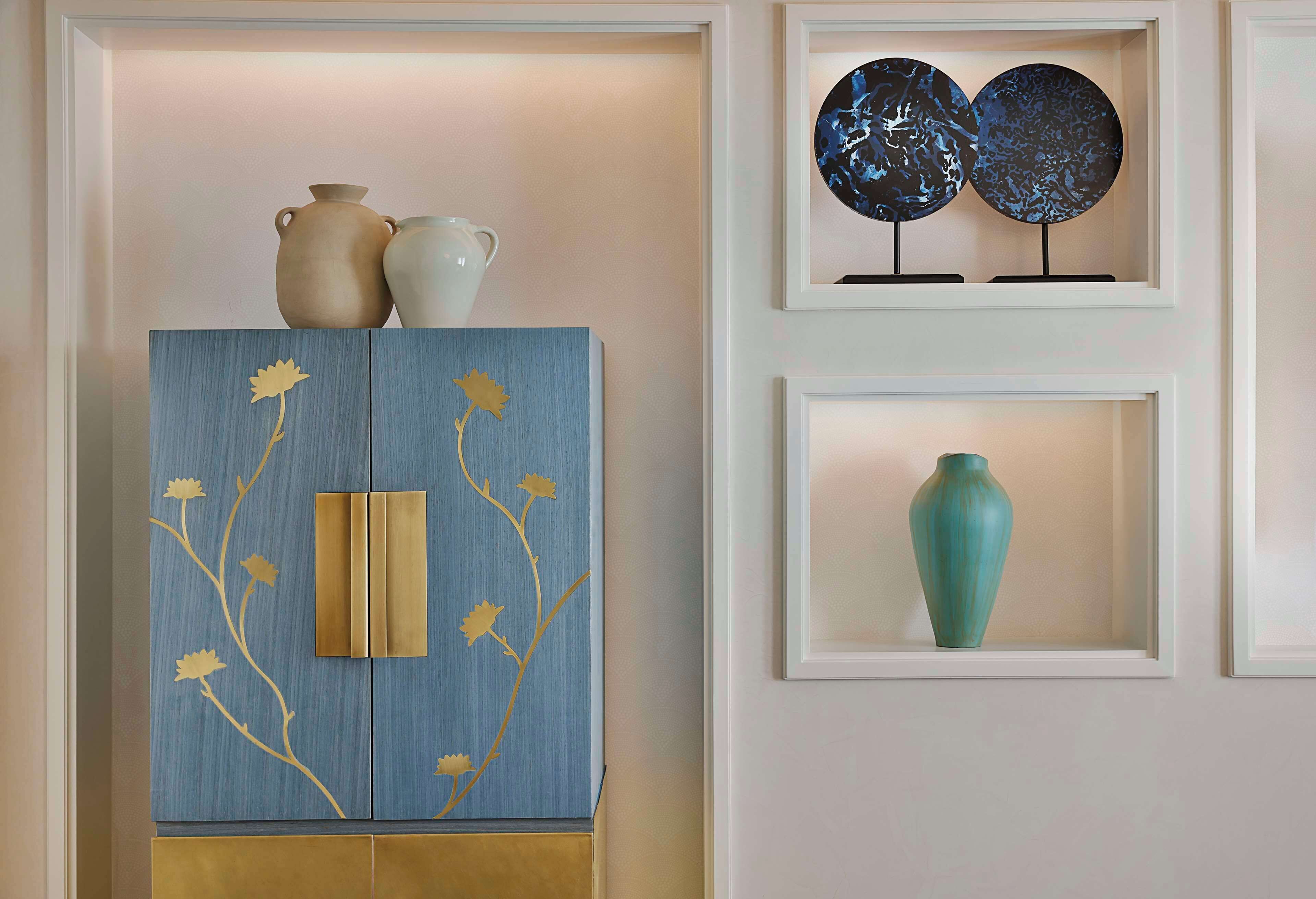 A blue cabinet with gold accents and floral designs, a white vase, and two abstract art pieces on the wall.