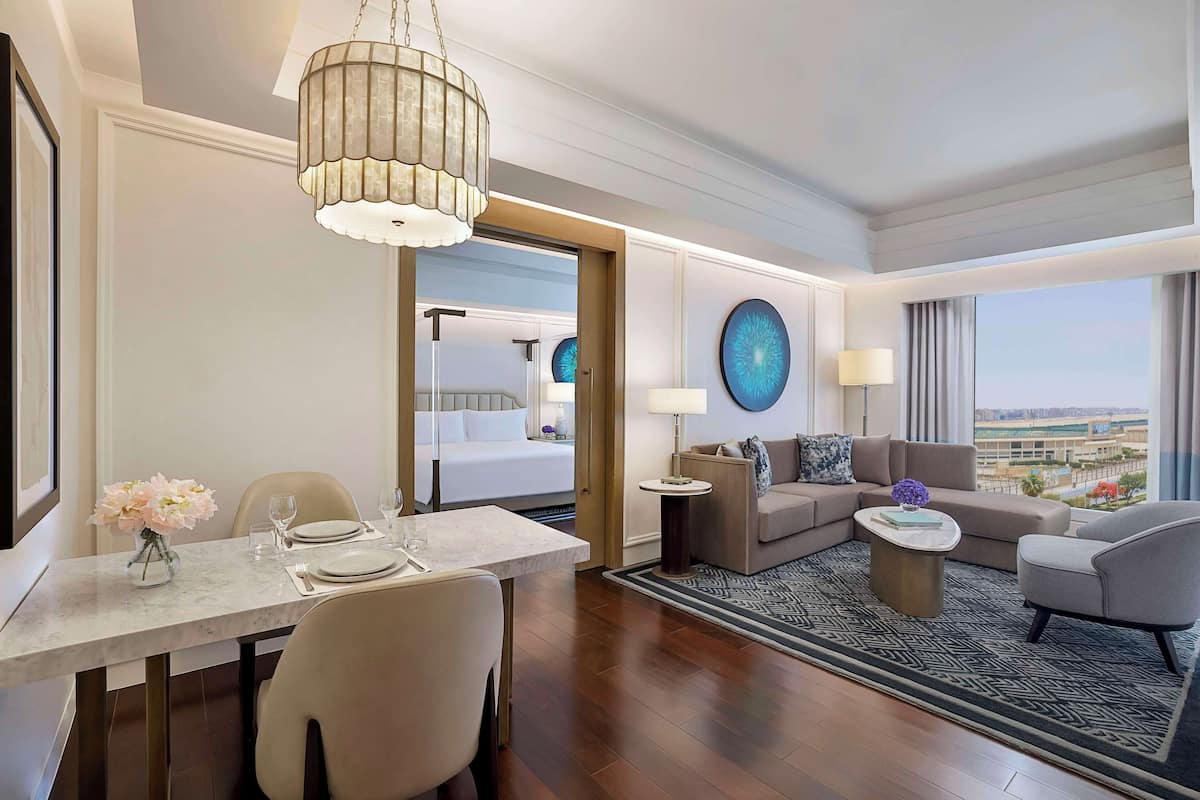 A modern hotel room with a dining area, a living room with a sofa and armchairs, a large window with a view, and a chandelier.
