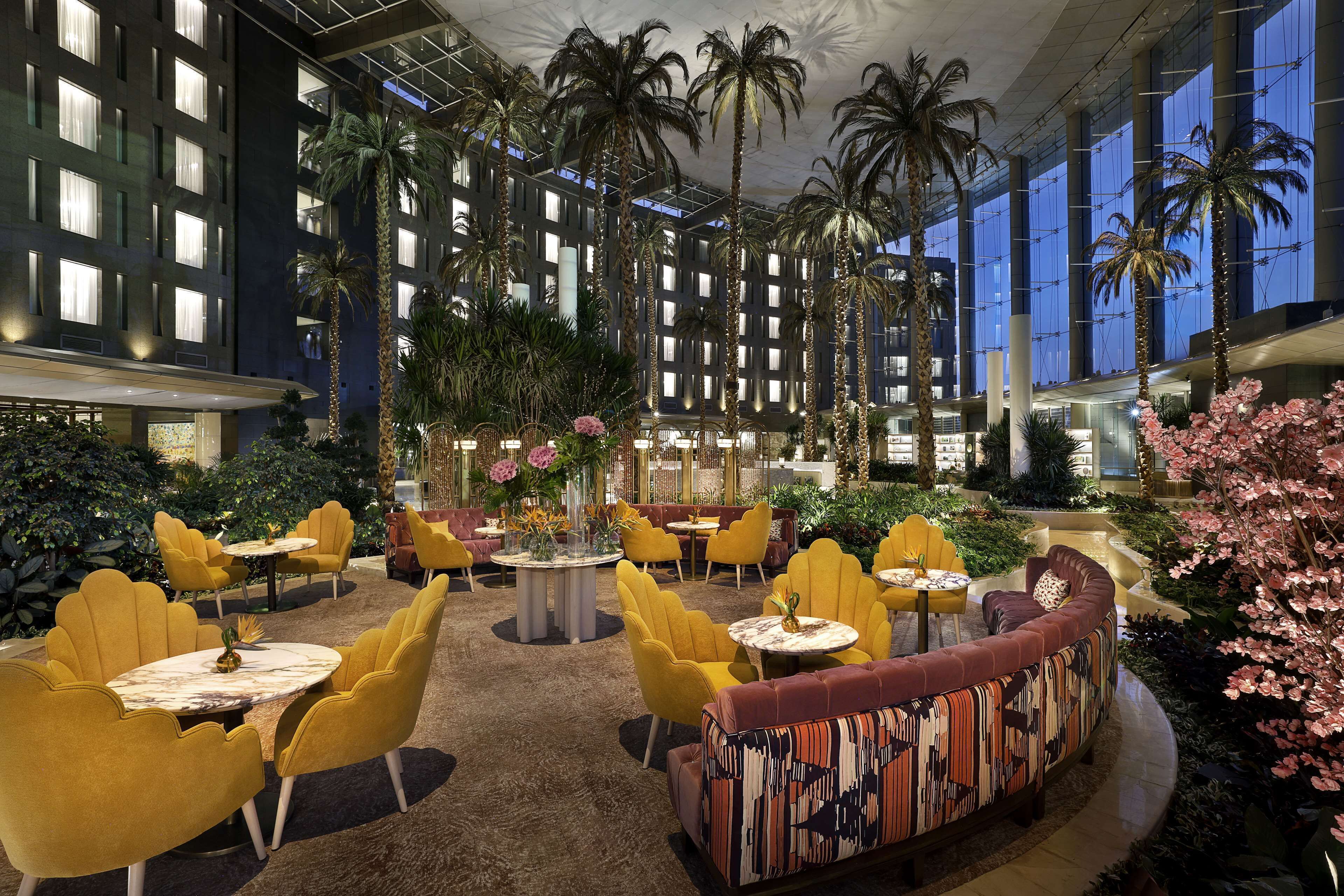 A spacious indoor atrium with high ceilings, palm trees, and yellow chairs around tables.
