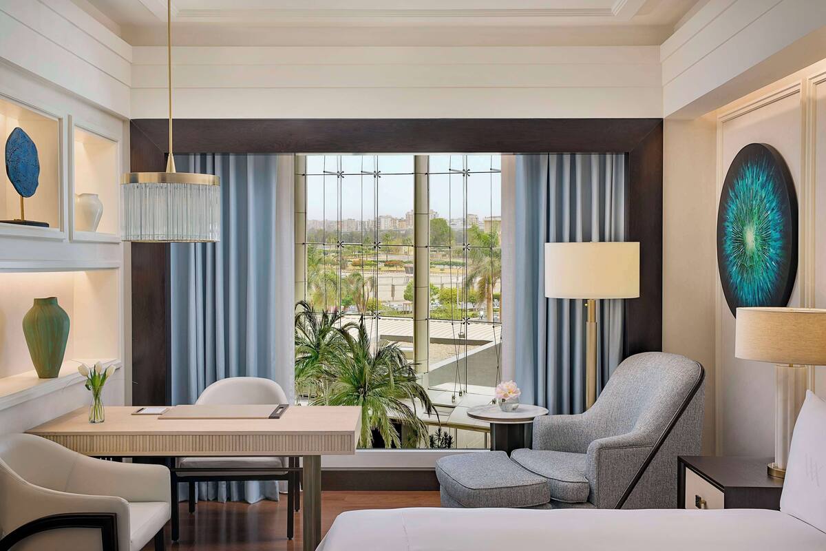 A hotel room with a bed, a desk, a chair, a small table, and a large window with curtains.