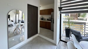 A modern hotel room with a large mirror, a dark door, a kitchen area with wooden cabinets, and a balcony with outdoor seating.