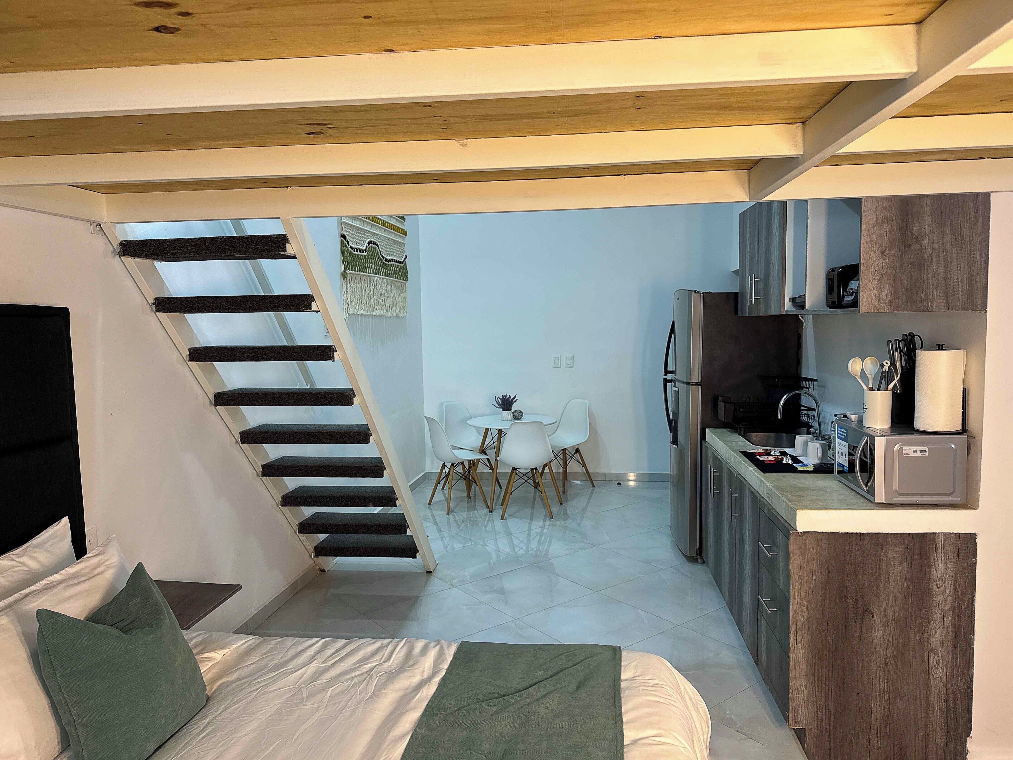 A modern bedroom with a bed, green pillows, a staircase, a dining area with a table and chairs, and a kitchen with appliances.