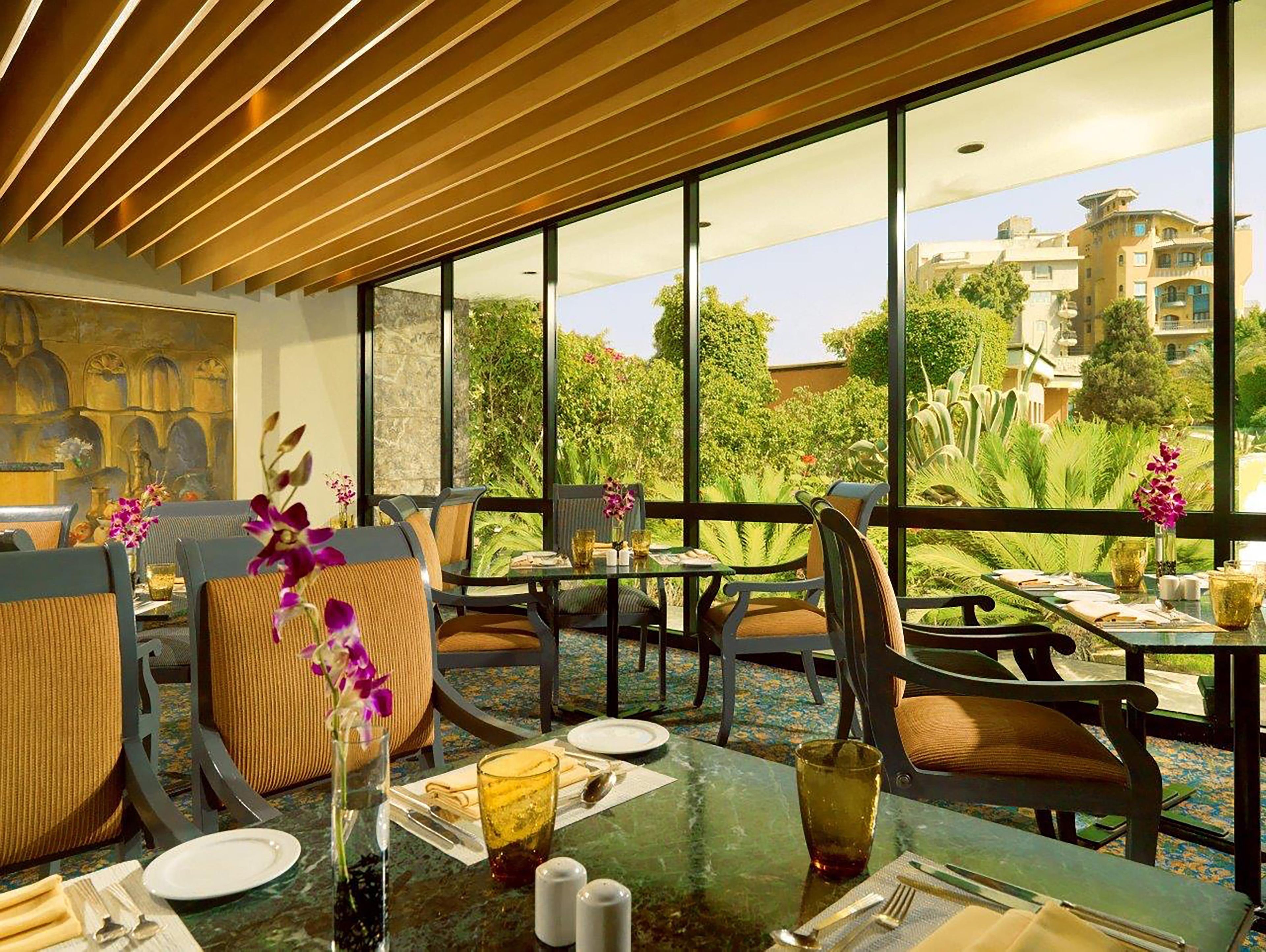 Zona pranzo con tavoli e sedie, ampie finestre con vista su spazi verdi ed edifici, e un soffitto con listelli di legno.
