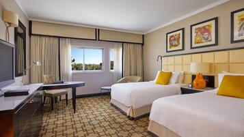Ein Hotelzimmer mit zwei Betten, einem Schreibtisch, einem Stuhl, einem Fenster mit Vorhängen und Bildern an der Wand.