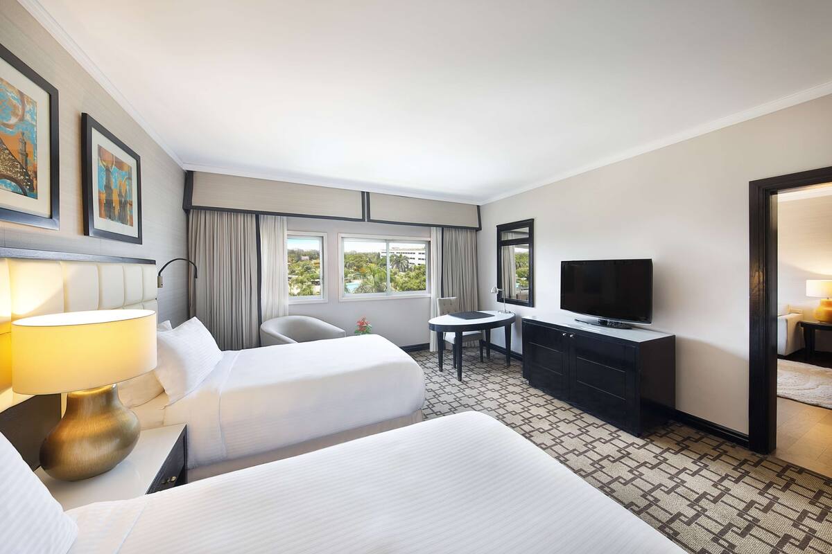 Quarto de hotel com duas camas, uma escrivaninha, uma televisão e janela com cortinas.