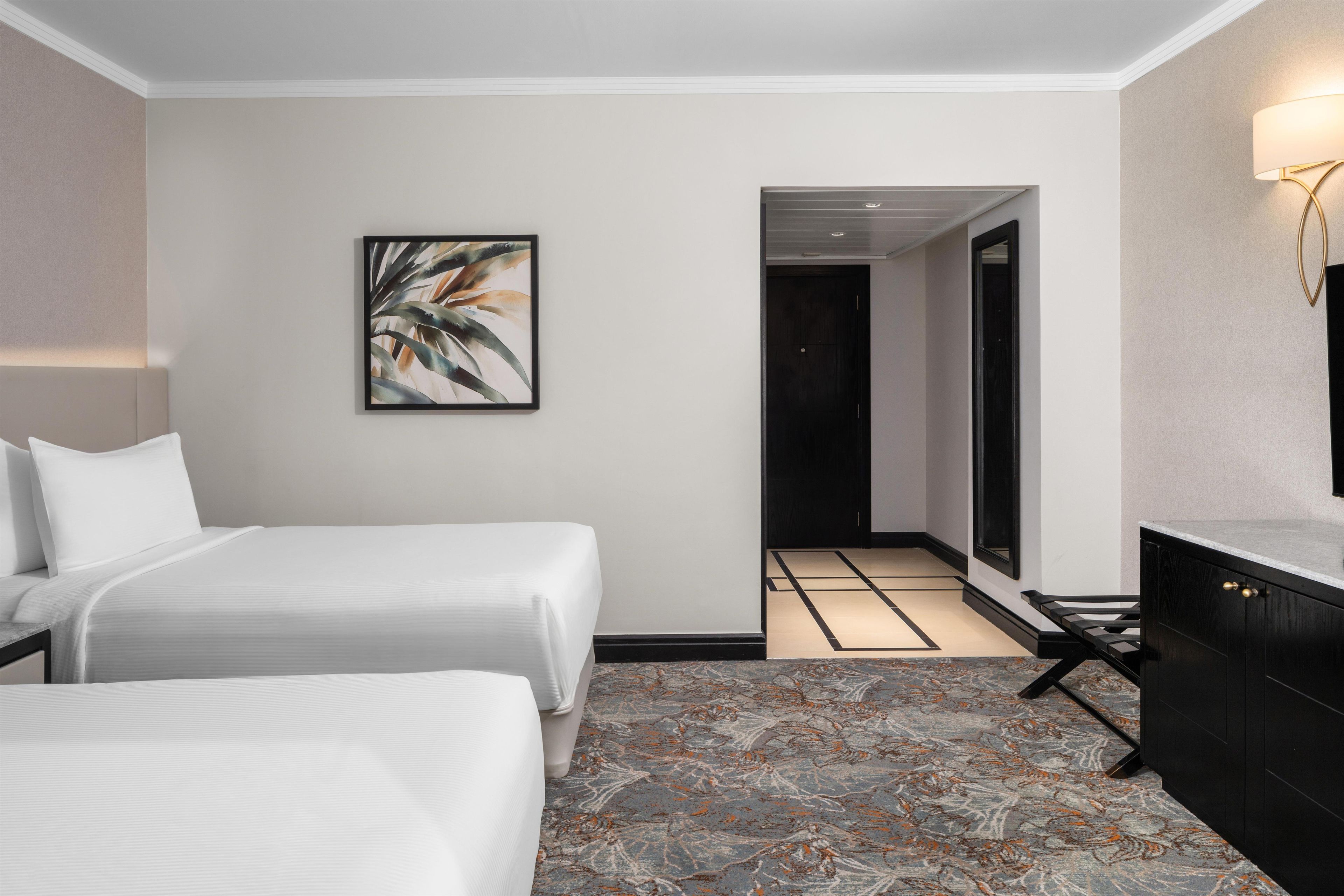 Ein Hotelzimmer mit zwei Betten, einem großen abstrakten Gemälde, einem modernen Badezimmer und einem Teppich mit Marmoroptik.