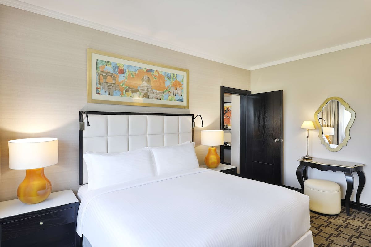 Quarto de hotel com uma cama grande, duas luminárias de cabeceira, uma mesinha de cabeceira, uma pequena mesa com abajur, um espelho e um quadro na parede.